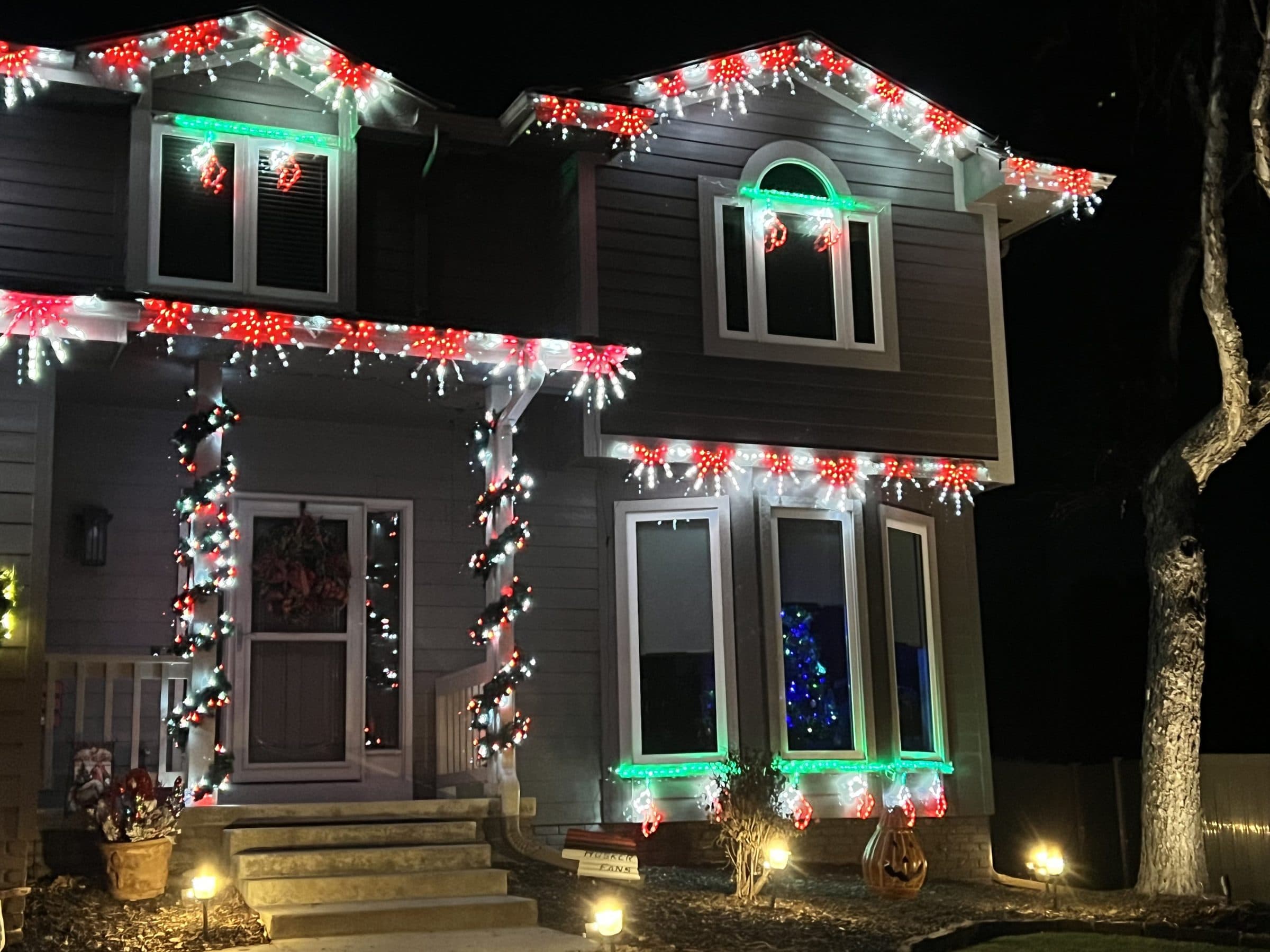 Front of a home with red and white lights, garland with red bows, and green lit windows