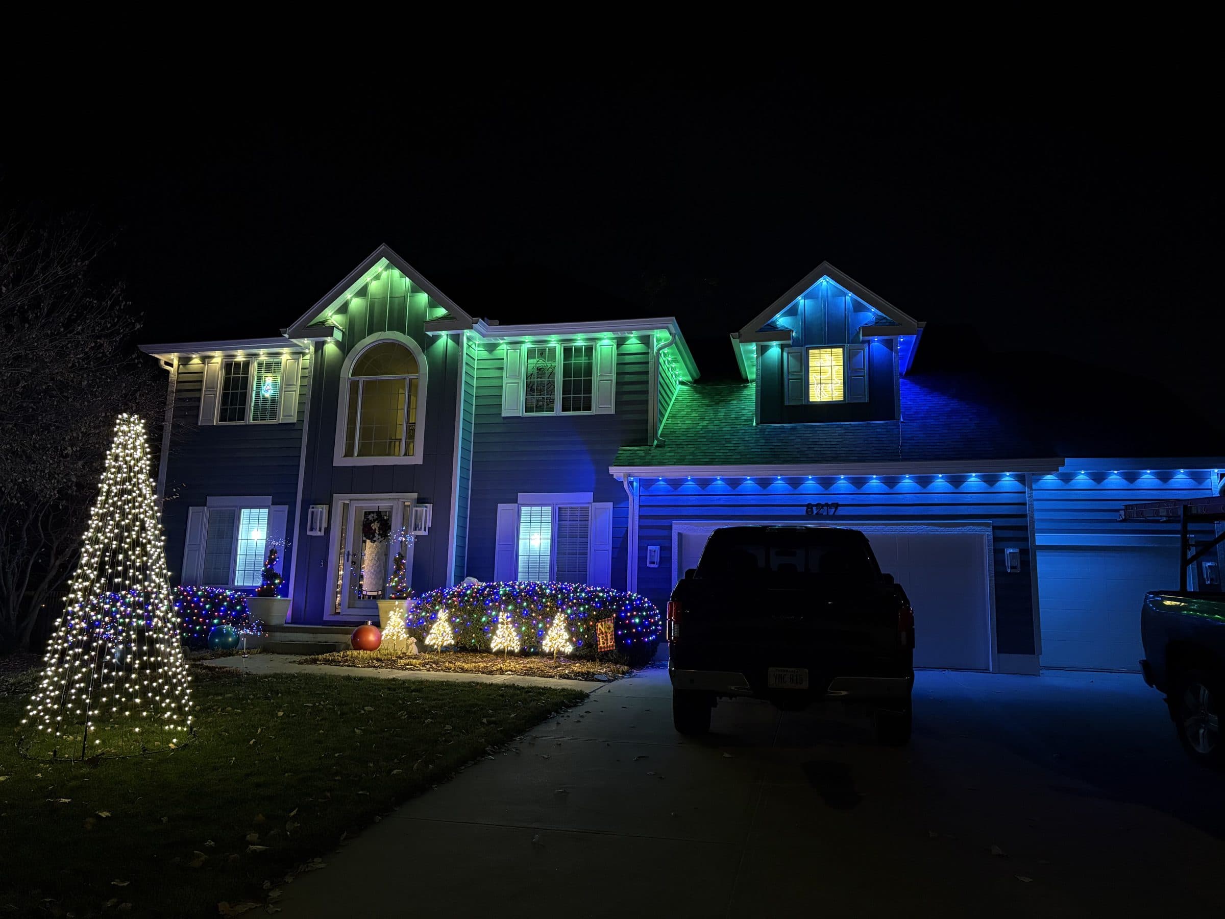 Two story home with green and blue roofline lights, lit shrubs and a lit Christmas tree in the yard